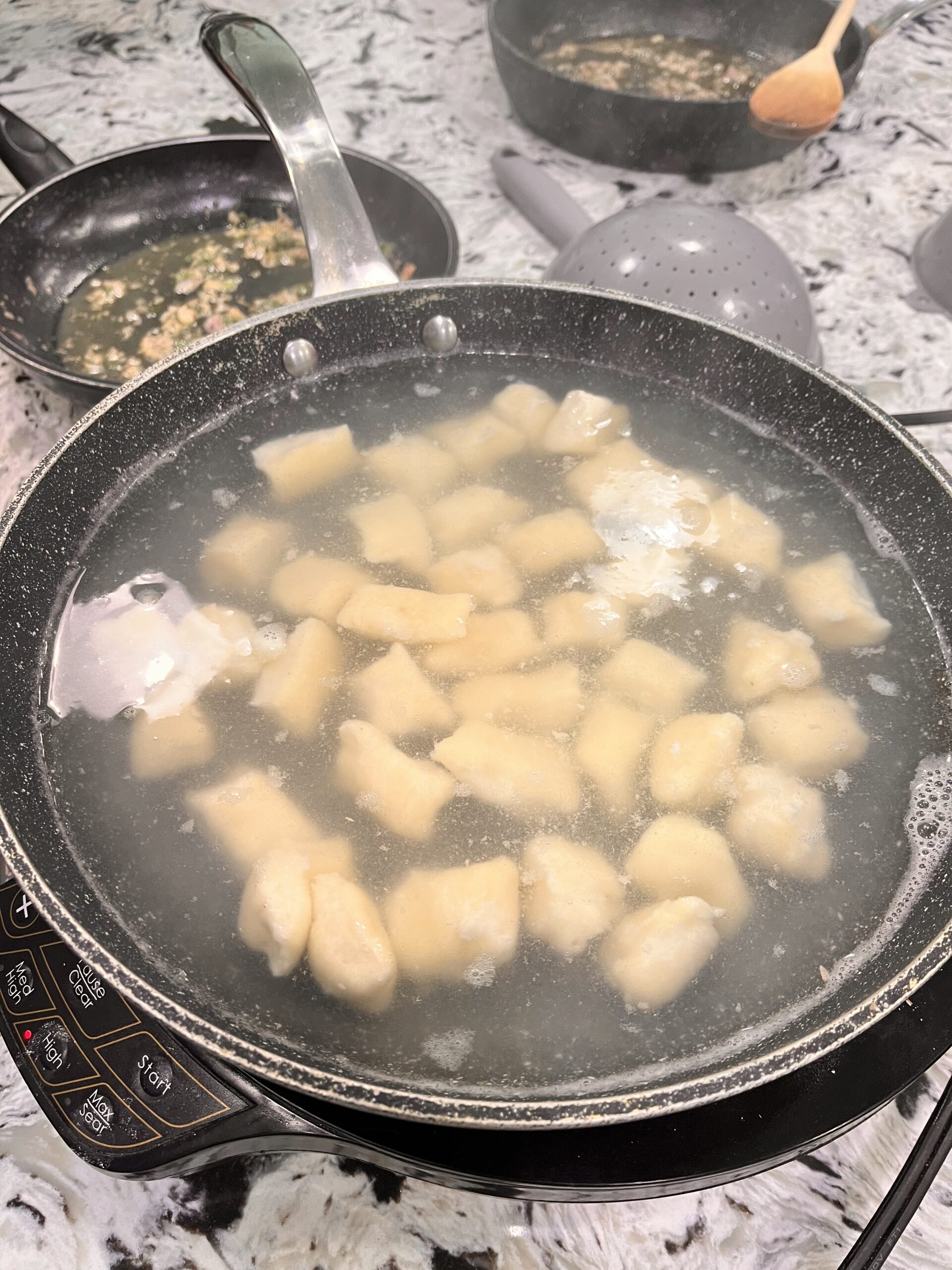 Frying pan filled with bitesize uncooked gnocchi dough and water set on a portable burner, sitting on a granite countertop. Two other frying pan and cooking spoons sit close by.