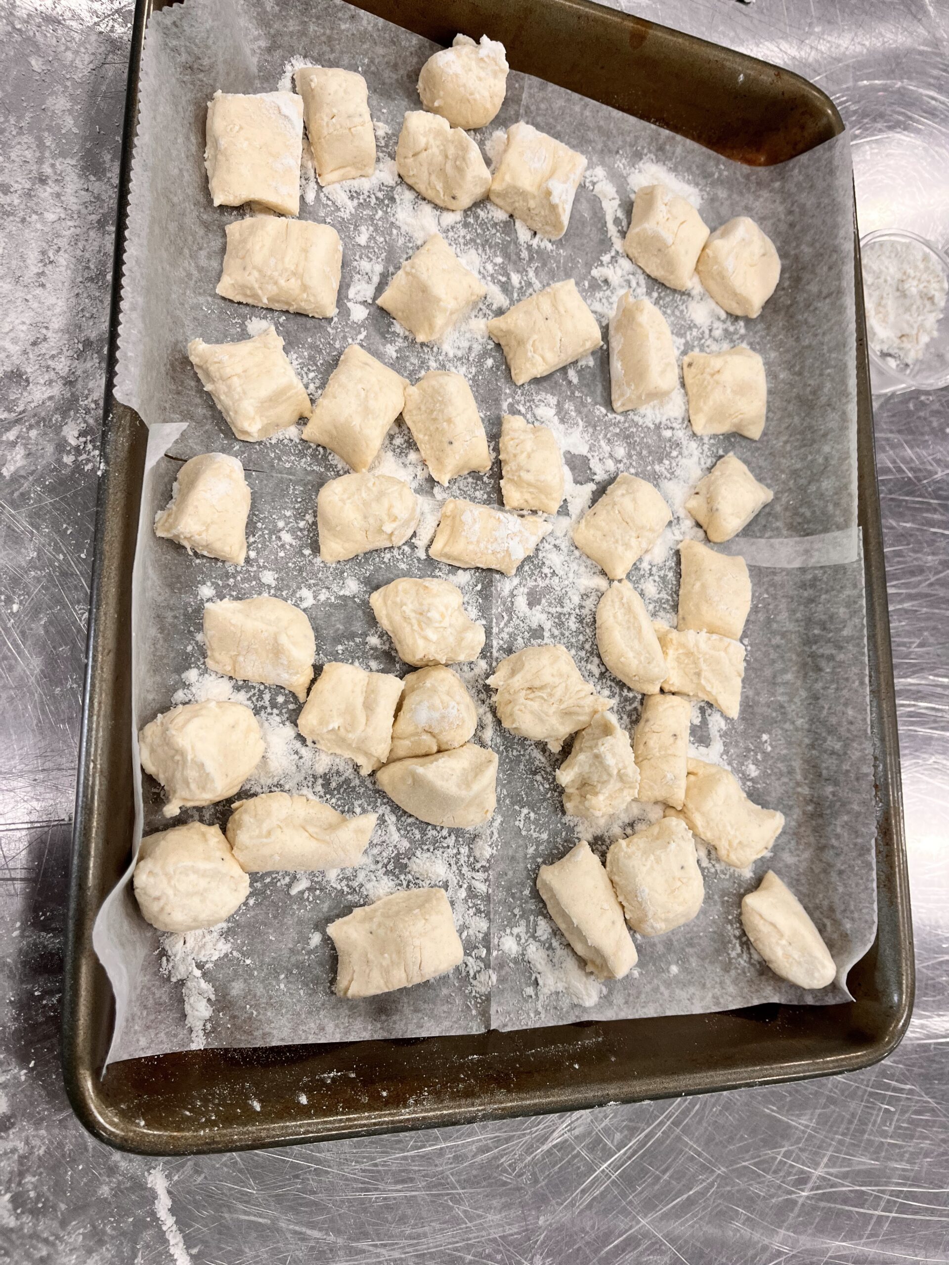 Baking pan lined with parchment paper filled with small bitesize uncooked gnocchi dough and sprinkles of flour.