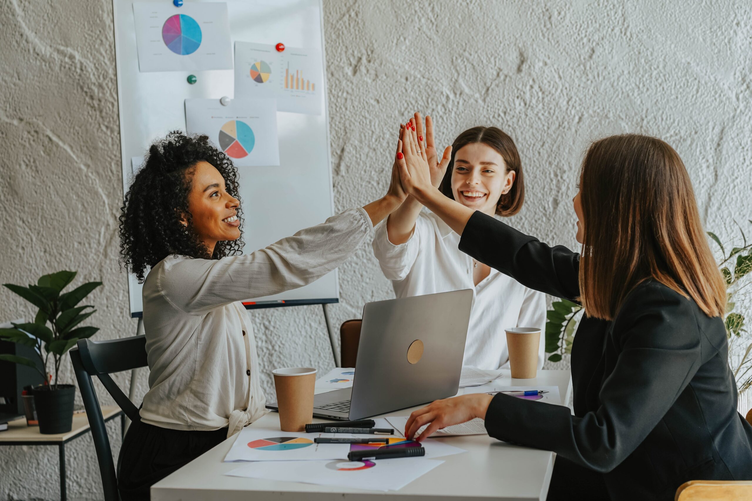 A group of 3 ladies sitting at a table with an open laptop, cups, writing utensils, and several pie charts giving each other a 3-way high-five.