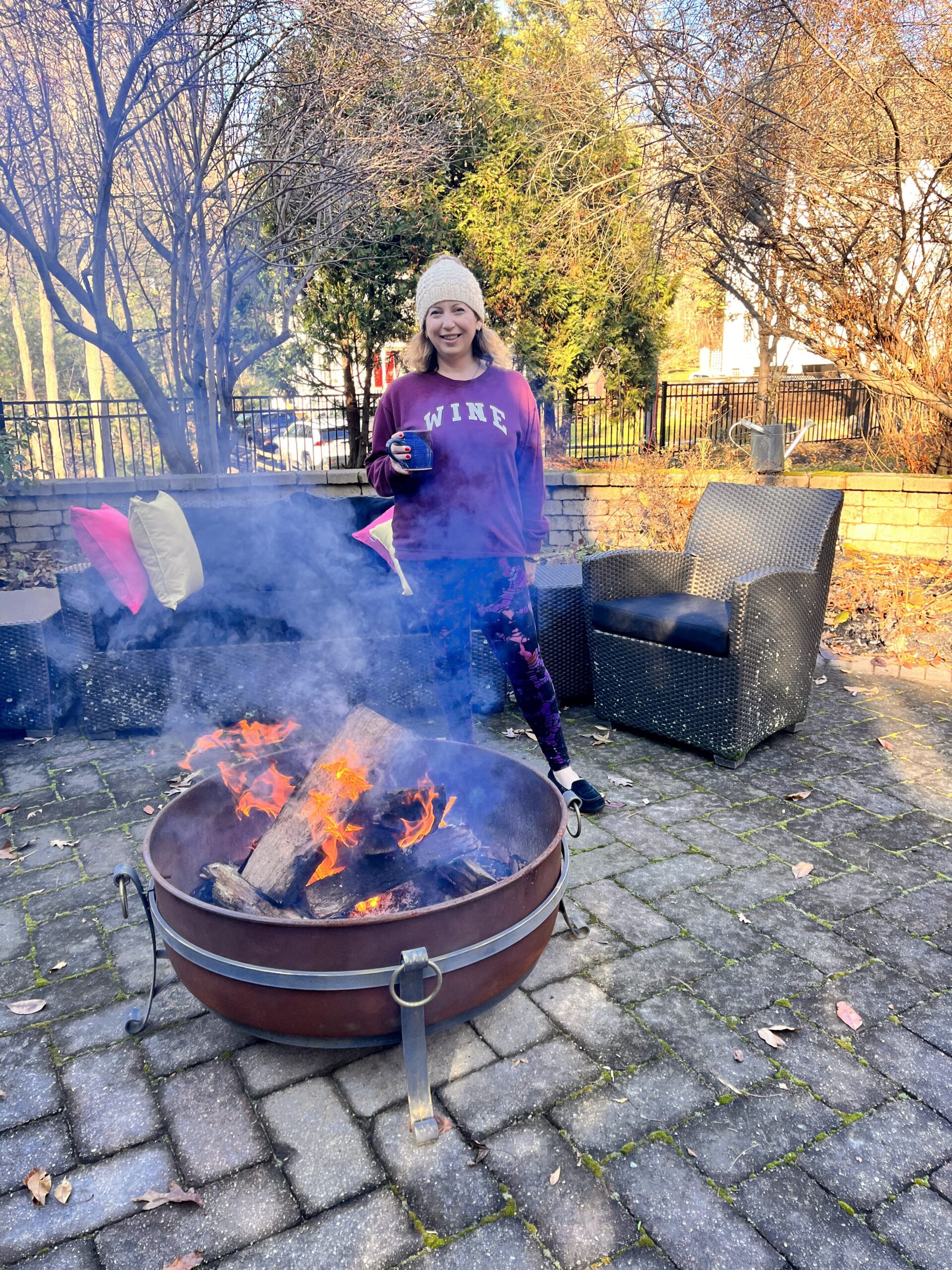 A woman stands next to a burning fire pit with smoke escaping the fire with an outdoor setting set behind her and a brick and metal fence and trees in the background.