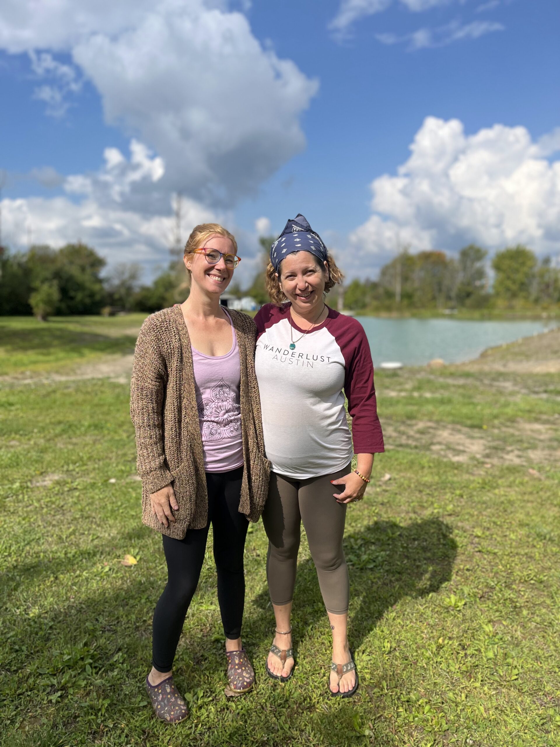 Two women stand close to one another in and open field smiling after completing a private yoga session.