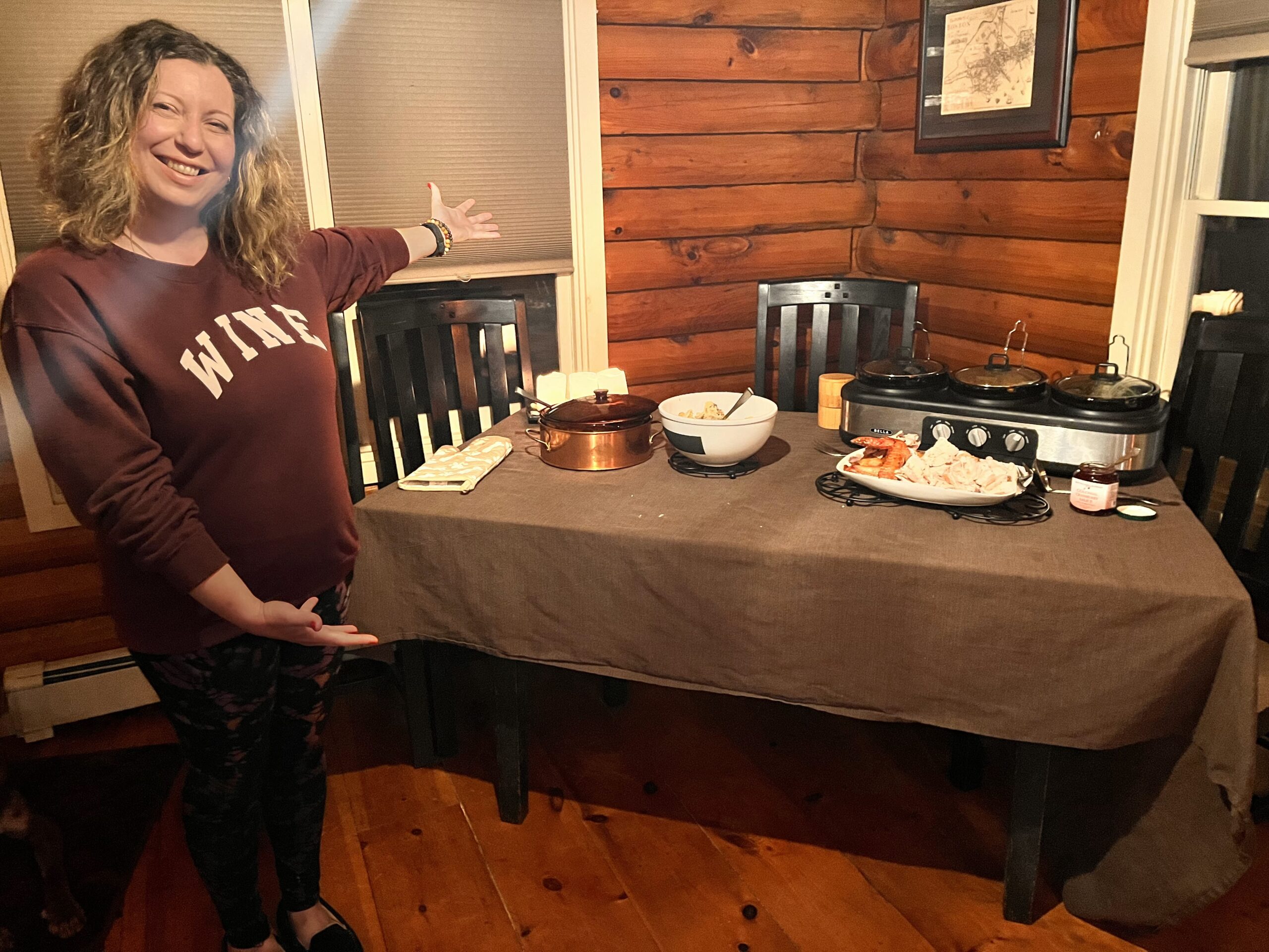 A woman stands next to a table covered with a brown table cloth. A three compartment food warmer and other containers filled with food occupy the tabletop. Three black wooden chairs are tucked in next to the table.