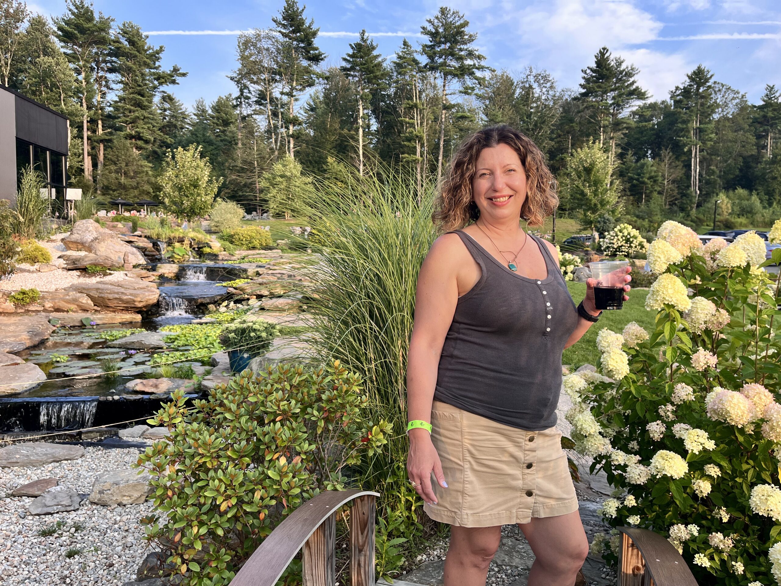 A woman holding a beer while standing next to a bush with yellow flowers on a bridge over a small creek.