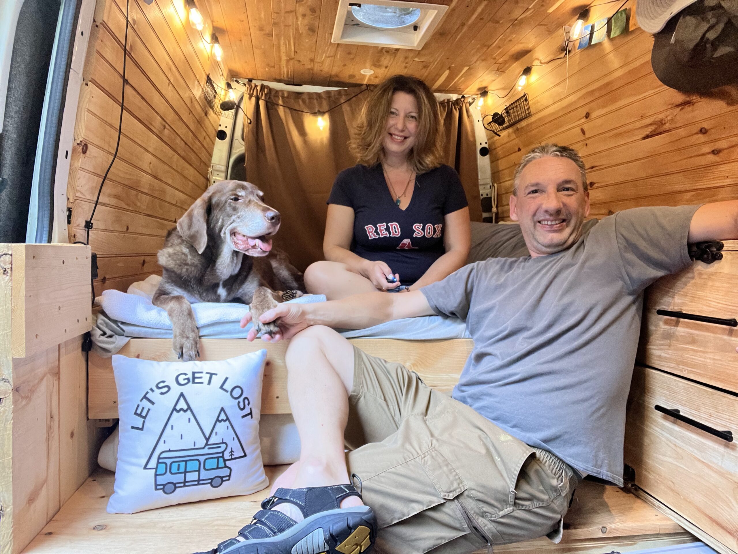 A woman, man and a dog in a wooden interior campervan with string lights all happy with a “Let’s get lost” pillow displayed.