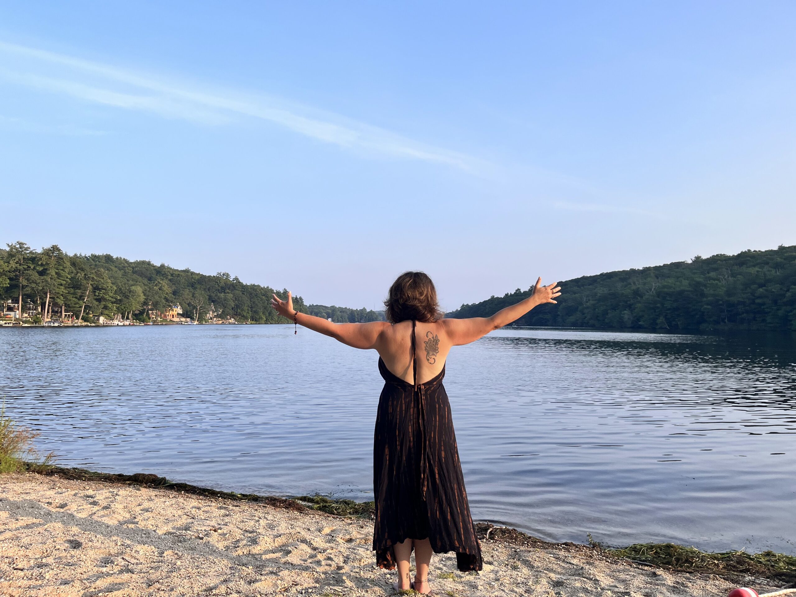 A woman confidently stands at the edge of a lake with outstretched arms in meditation overlooking the water.