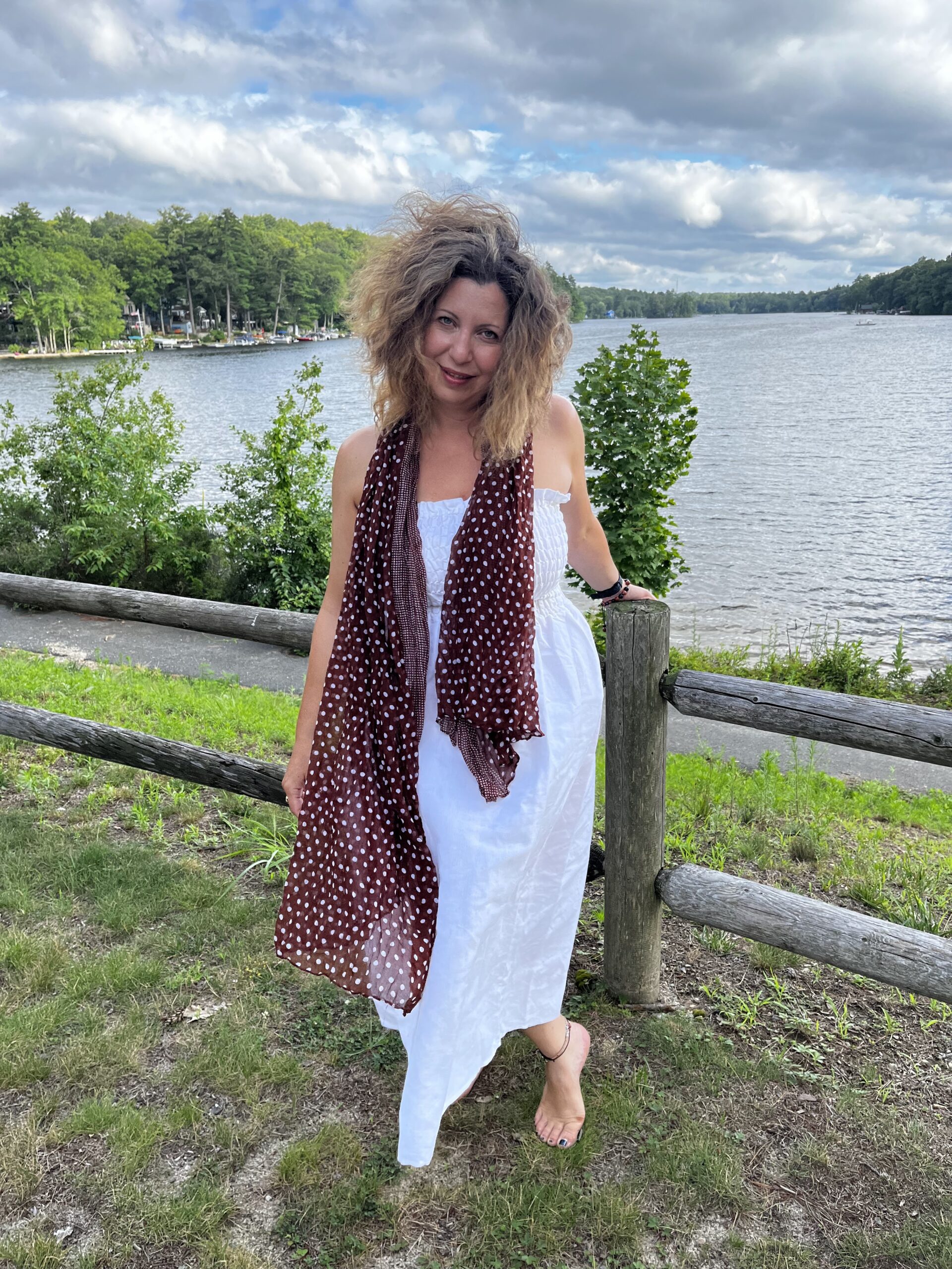 Woman wearing white dress and brown polka dot scarf holding a waist-height wooden fence in front of a lake. Tall and short green leafy trees surrounding lake.