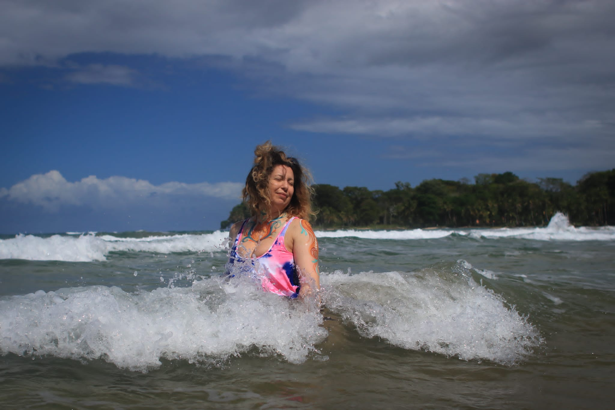 A woman sits on the beach as the waves crashes over her, under a partially cloudy blue sky.