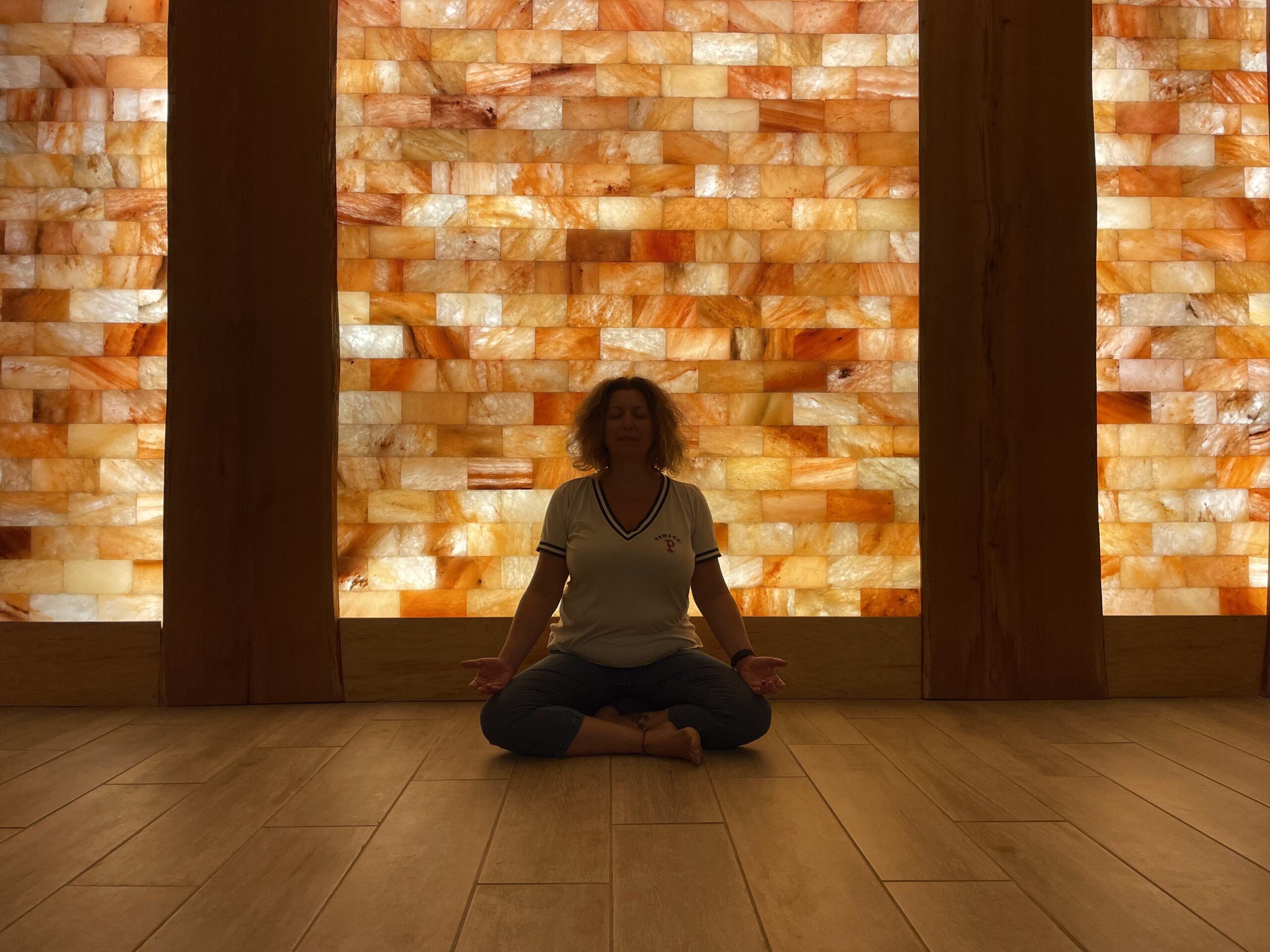 Woman in white shirt and black yoga pants sitting cross legged with hands on each knee slightly turned upwards. She is sitting on a wooden floor in room with low lighting and a wall with arranged with tiles of different shades of cream, orange, and brown. She is sitting in a salt room.