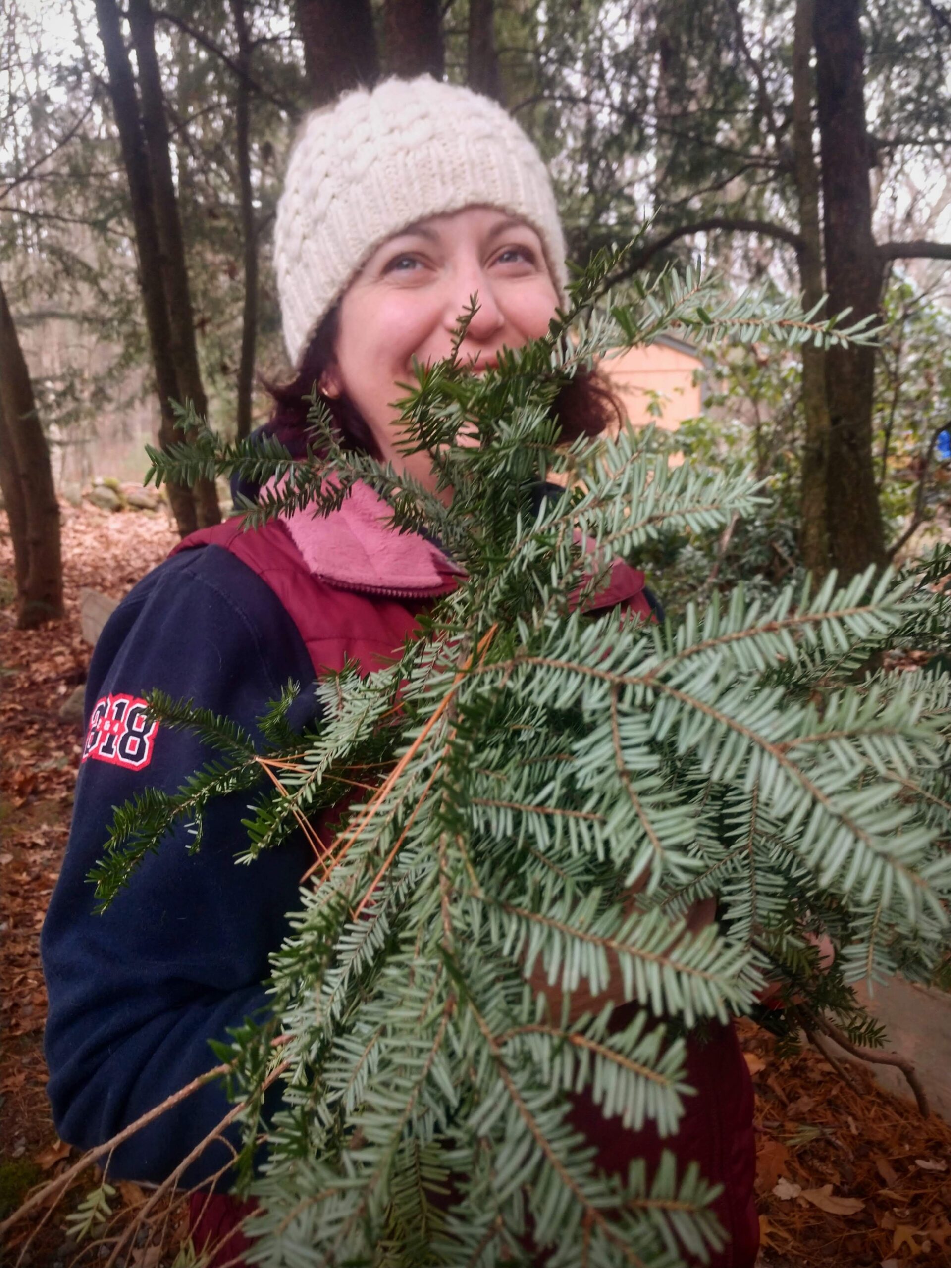 A woman dressed in a navy blue long sleeve shirt and a red vest over it and wearing a white knit beanie stands in the woods with pine leaves covering part of her face and body.