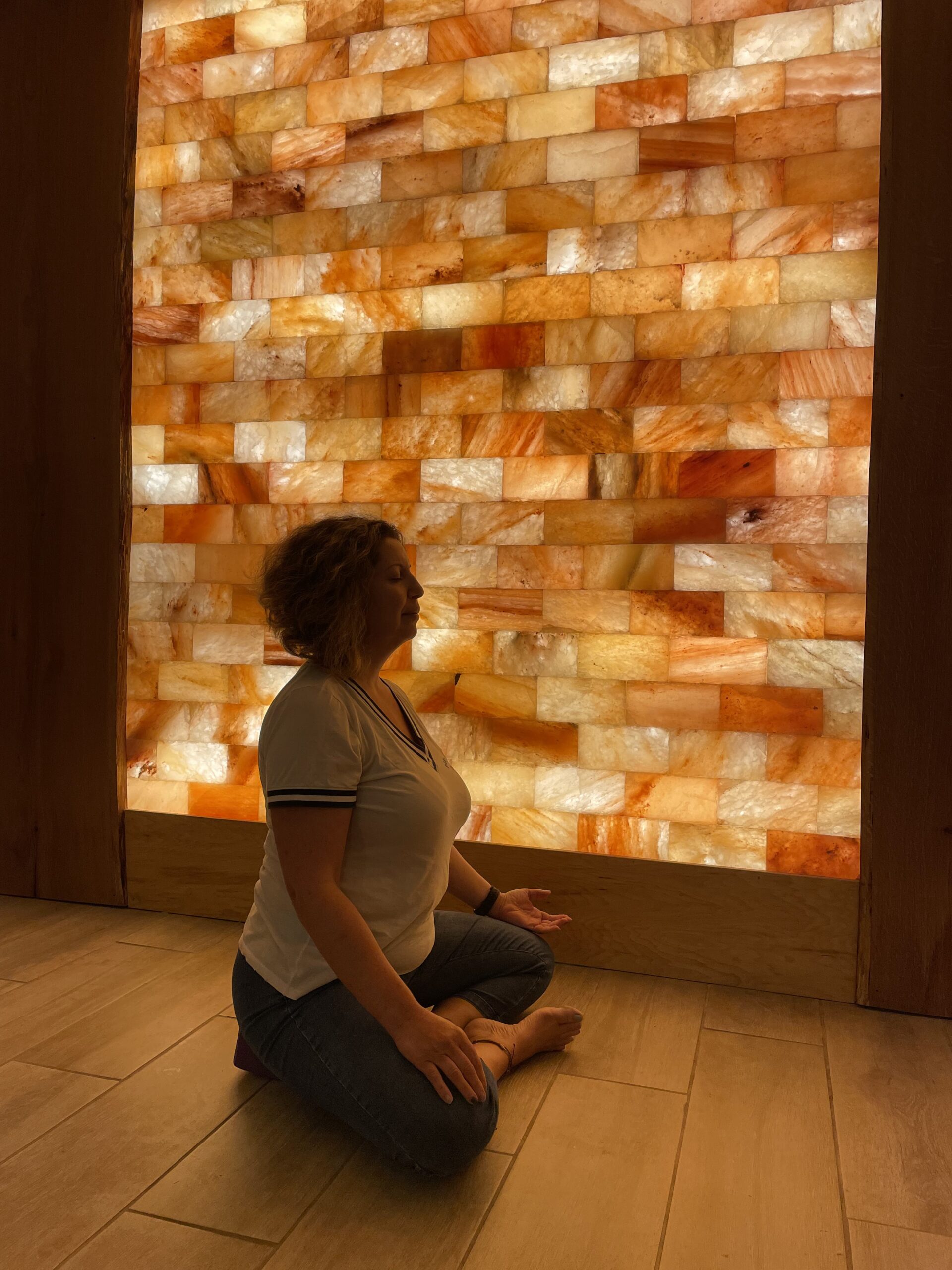 A woman in a meditative state sitting criss crossed on a wooden floor in the Just Breathe Salt Room with a lightly illuminated wall made up of tiles in different shades of brown.