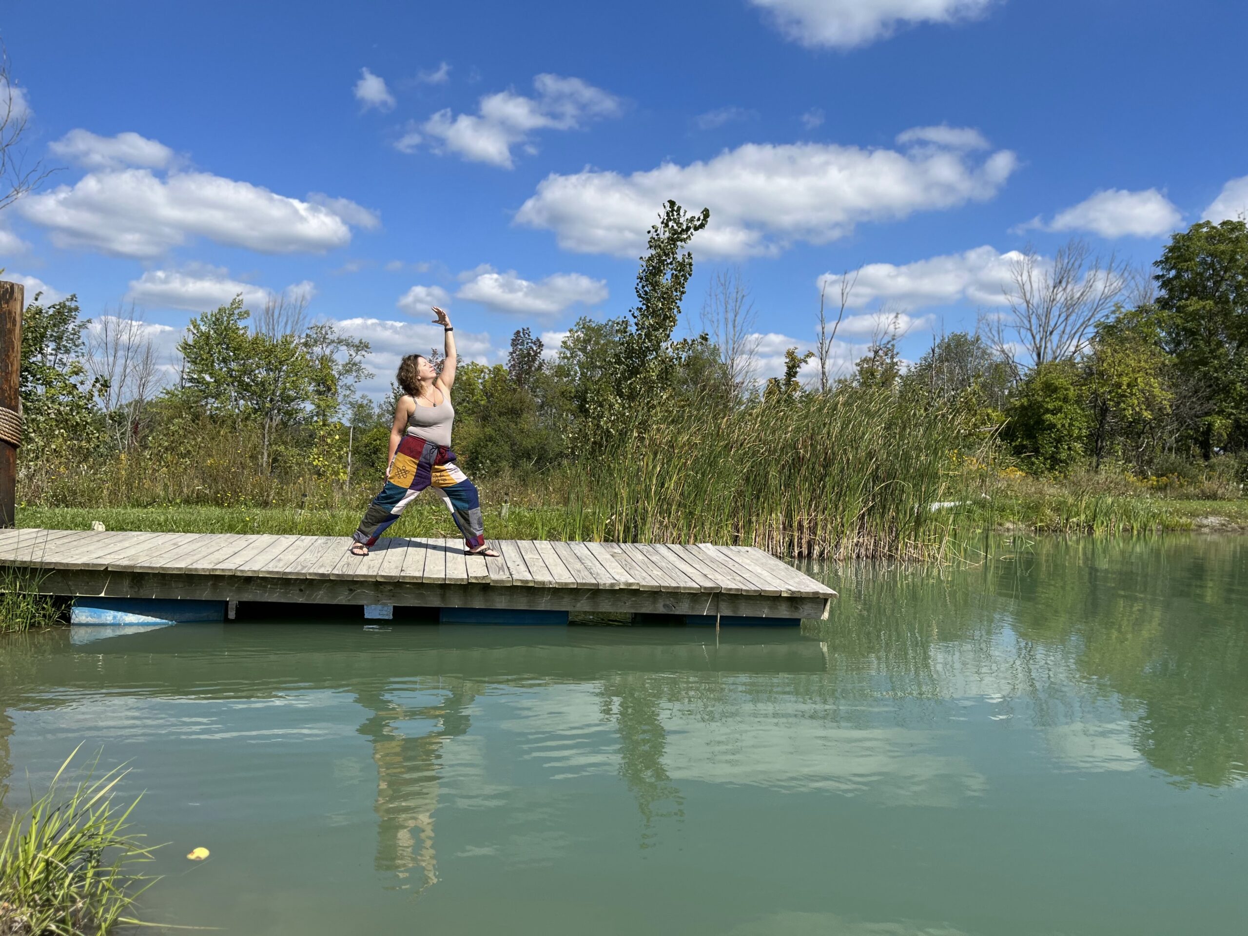 Woman in colorful patch-work pants and gray spaghetti top doing a warrior pose with left arm stretched upwards on a wooden dock over a pond with partly cloudy blue skies and tall green trees in the background.