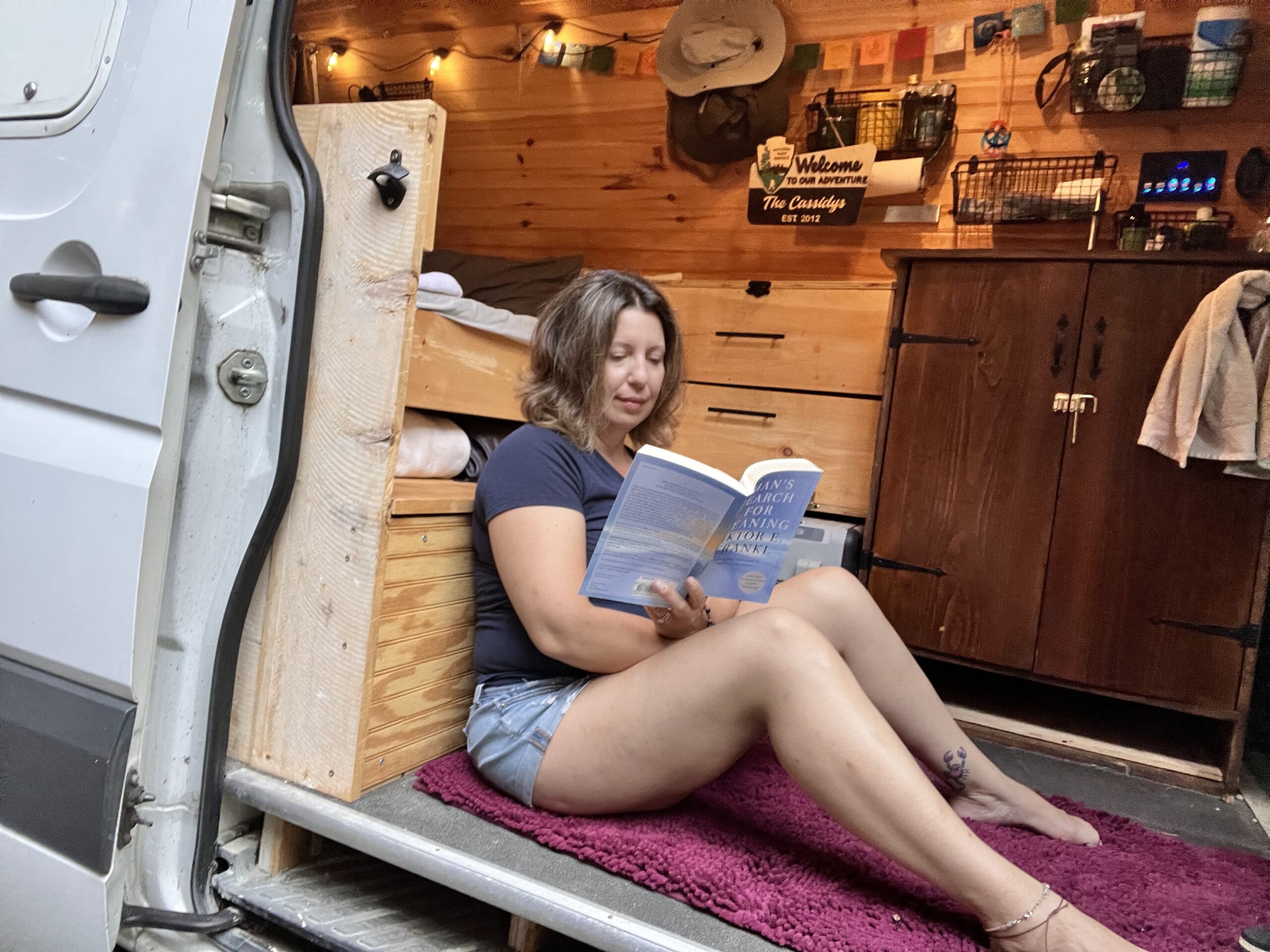 A woman peacefully sits on the floor of her campervan reading a book.