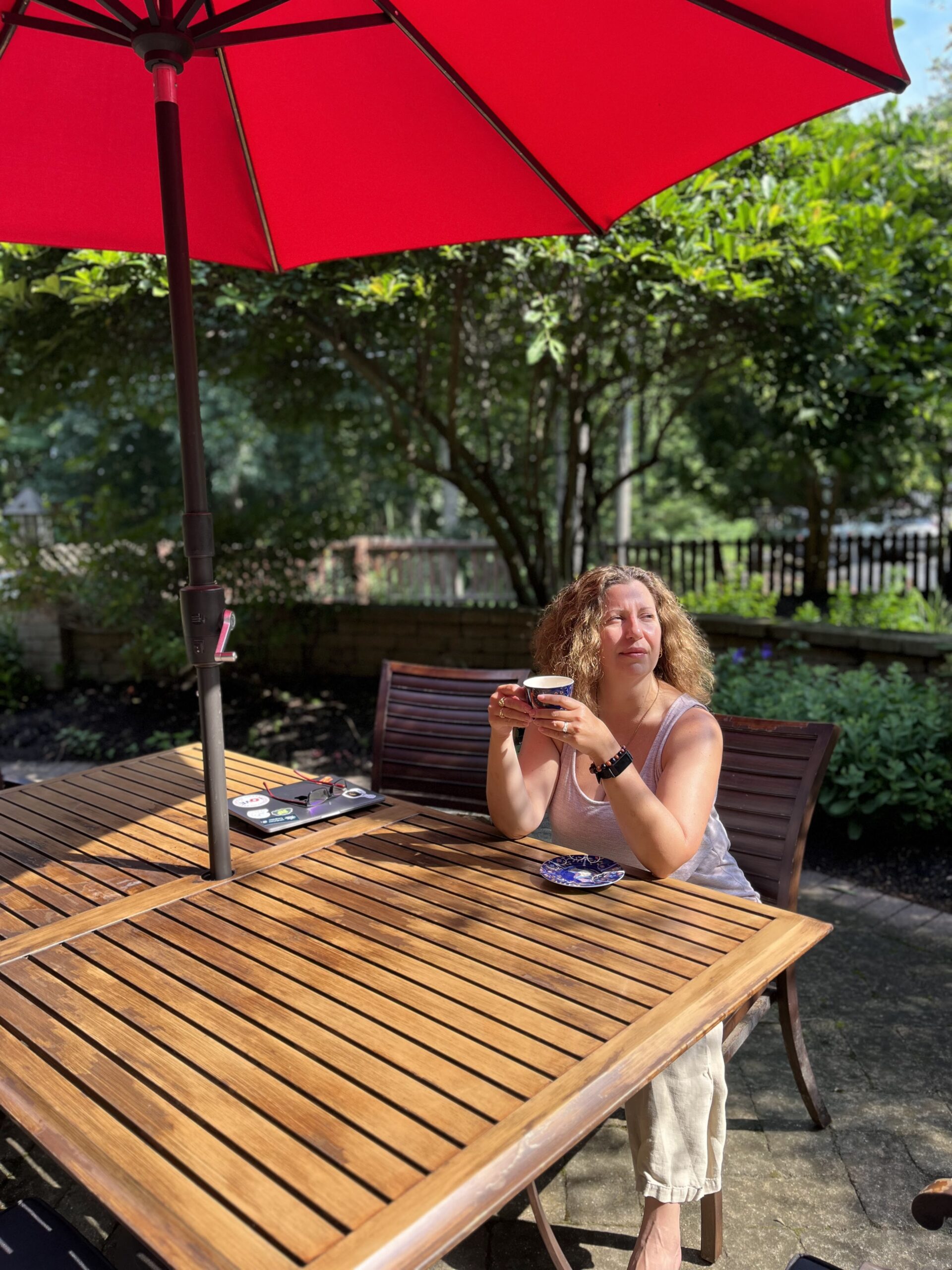 A blond woman sits in a meditative state at a table under an umbrella in her backyard sipping on a hot drink as she gazes in the distance.