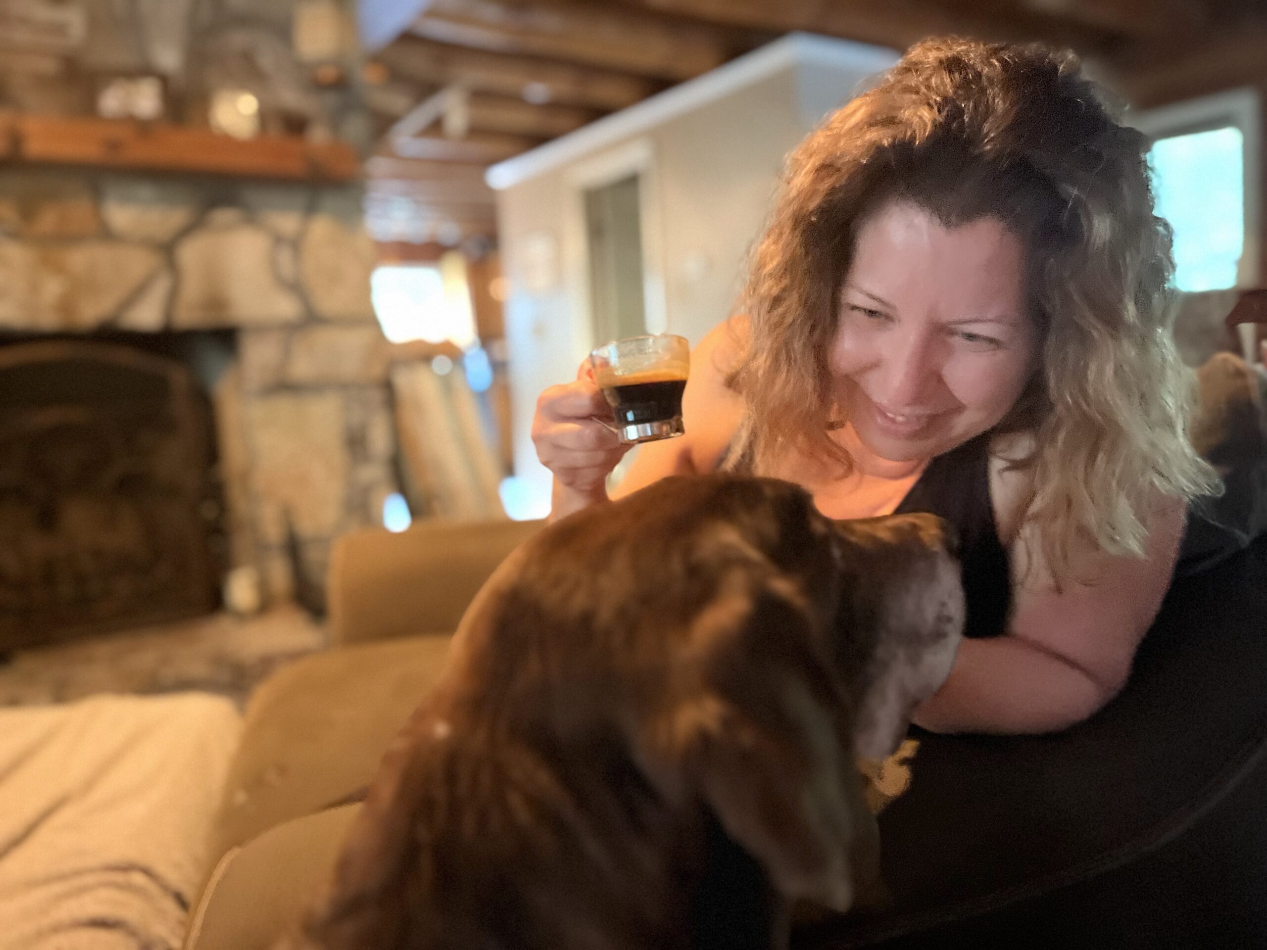 A woman happily gazes at her pup while holding an espresso on her couch in her living room.