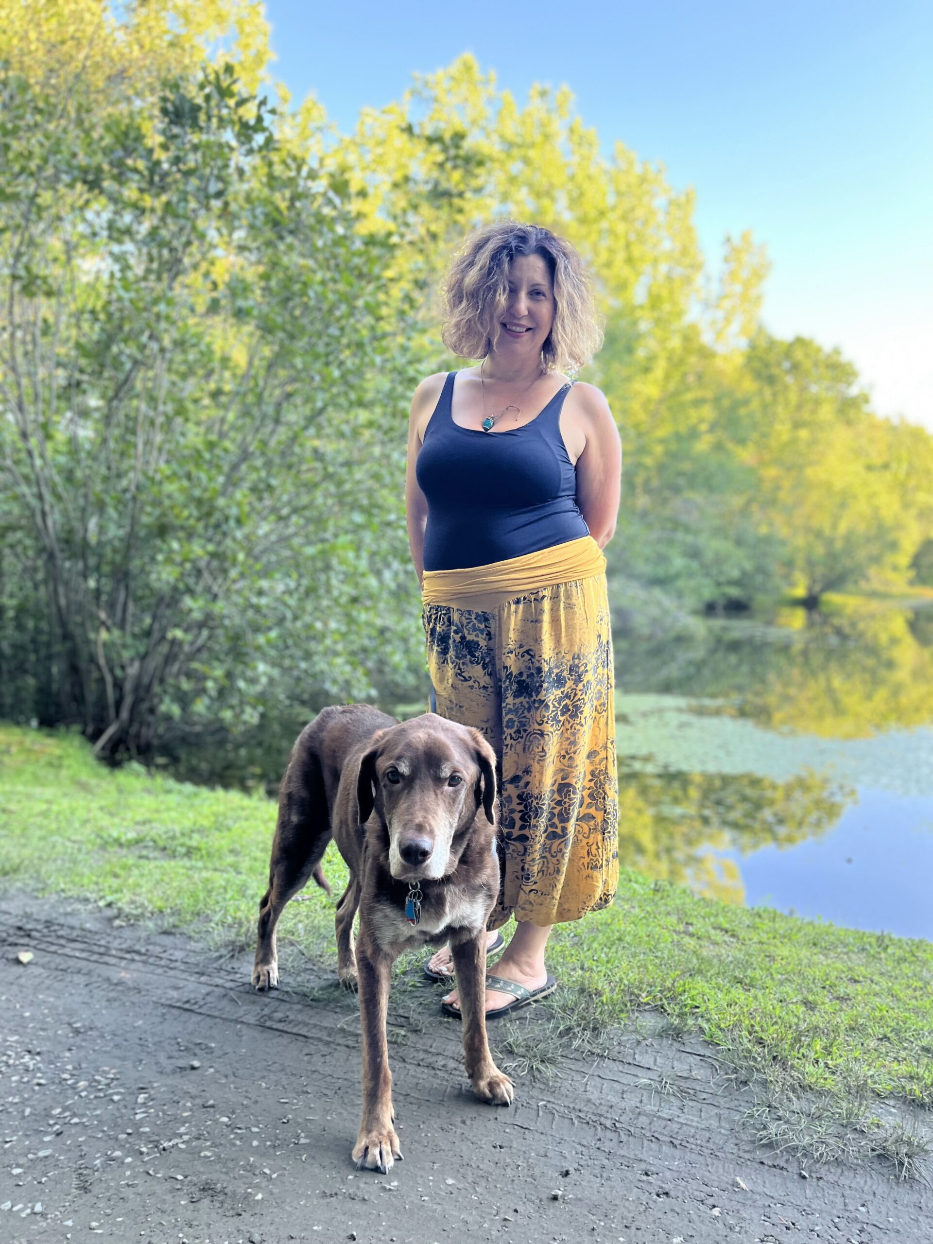 Woman in navy blue tank top with yellow flowery pants on a walking path by a lake in the woods with her white and brown dog.