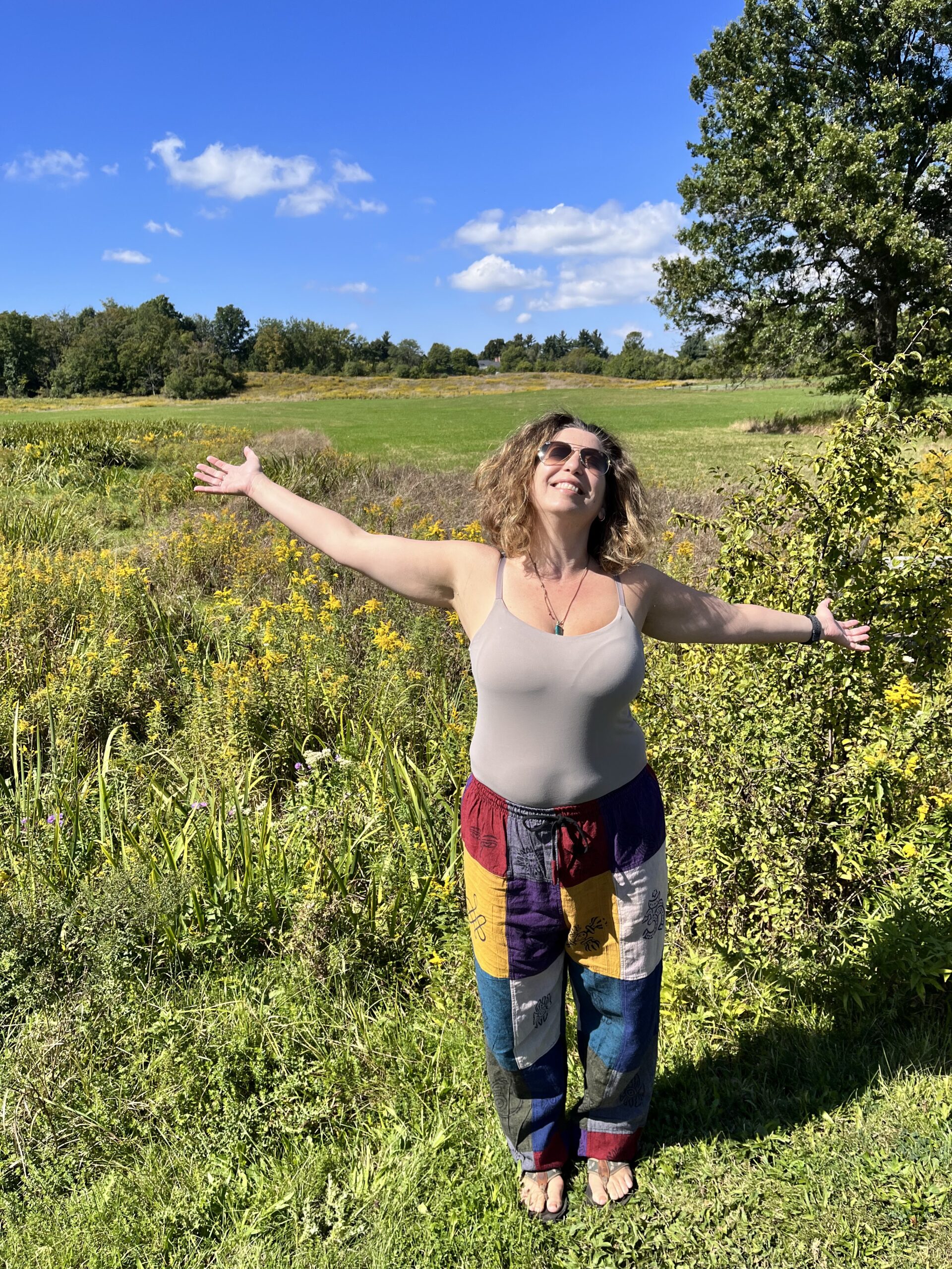 Woman with arms open wide wearing sunglasses, gray spaghetti strap shirt, and colorful patchwork pants looking up into the sky on a sunny day, soaking in the sunshine, with partially cloudy blue sky. She is standing on green grass with a bush of yellow flowers behind her, tall green trees, and an open field in the background.