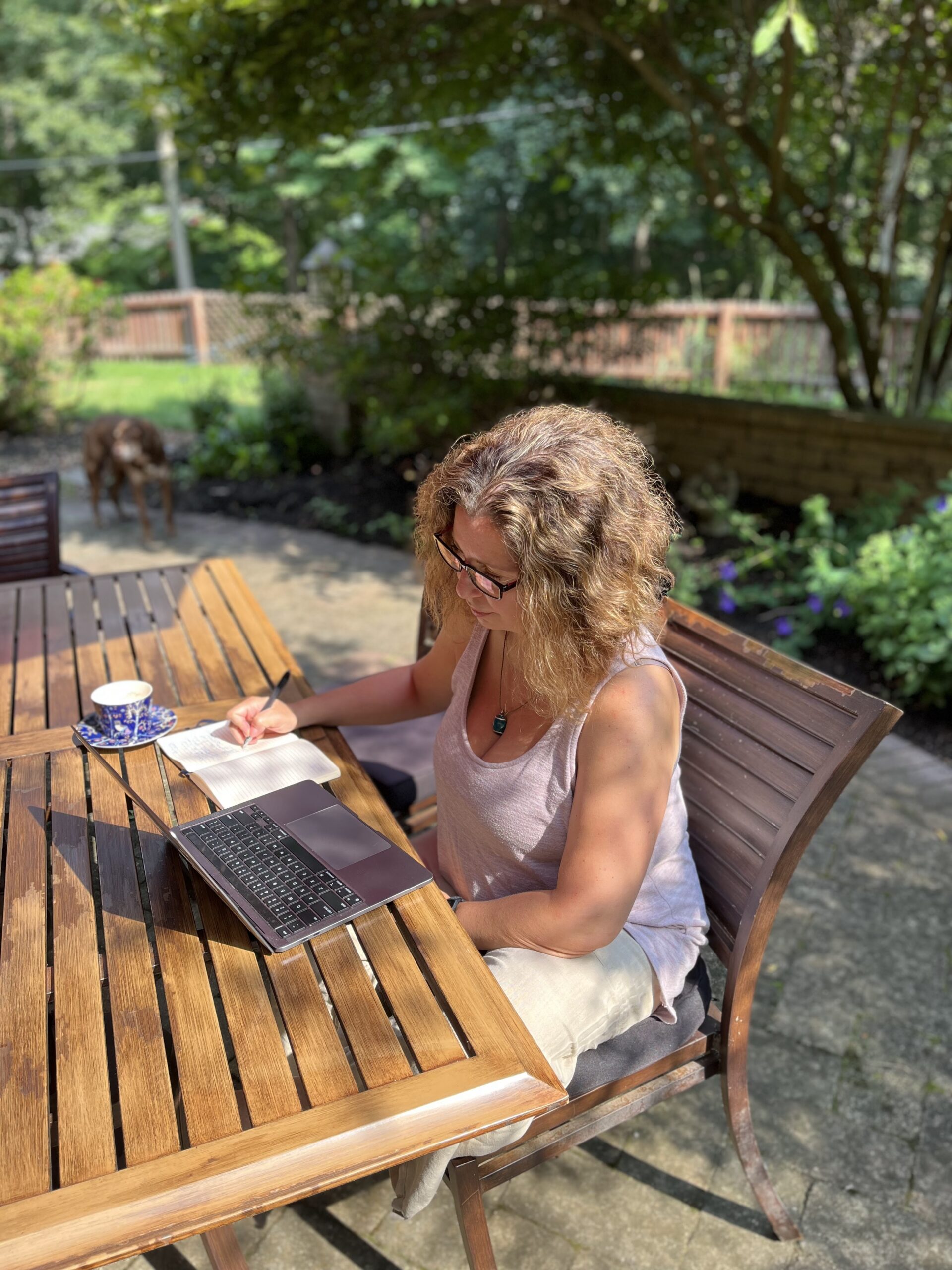 Woman in her garden sitting at a wooden table holding a pen and writing in a notebook with a cup of tea and an open laptop.