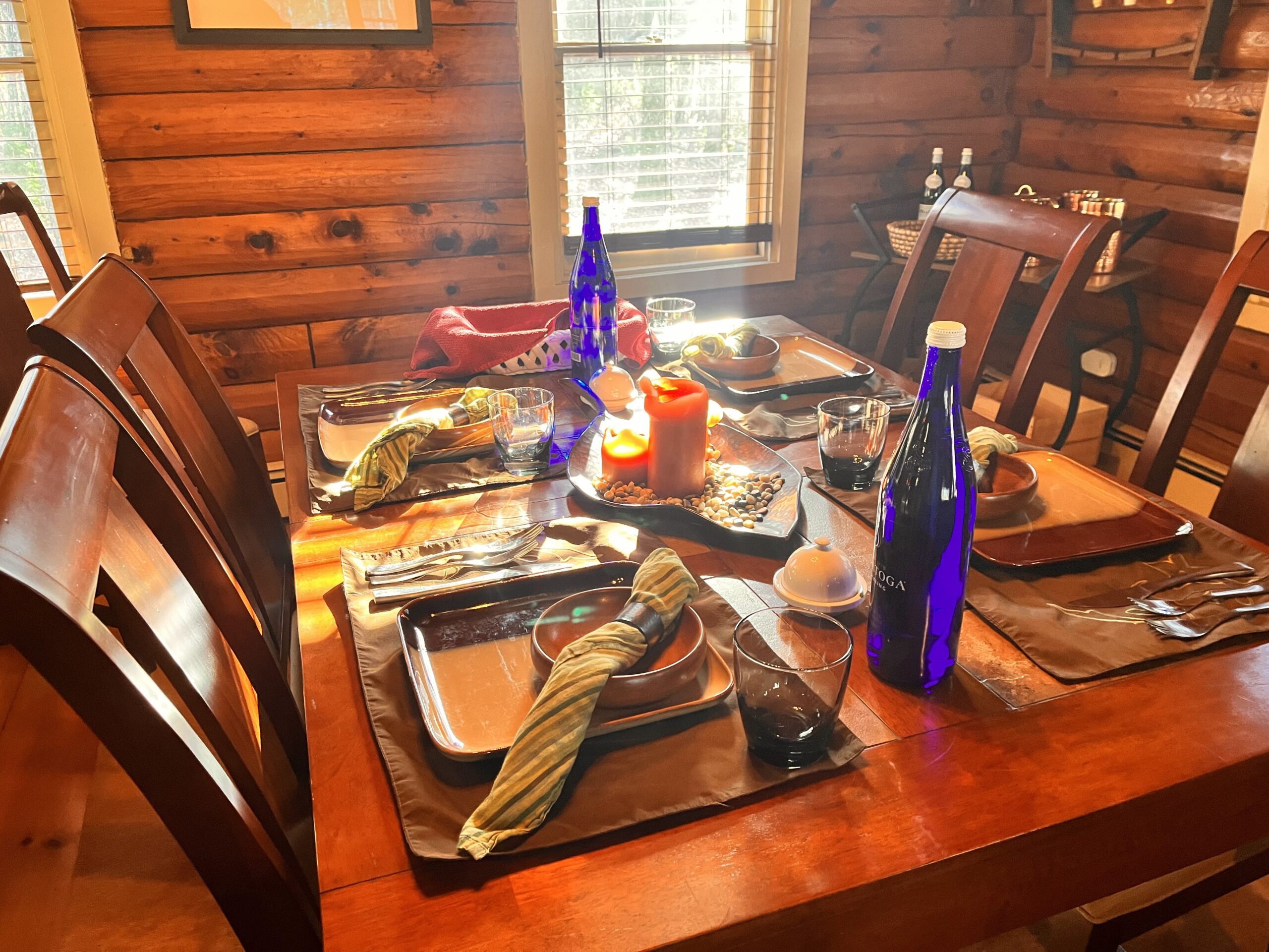 A table set for 4, where each place setting has a placemat, a square ceramic plate, a small ceramic bowl, a glass cup, 2 forks, a butter knife, and a napkin with a ring around it. The table also has two large blue bottles of water and a centerpiece with two candles.