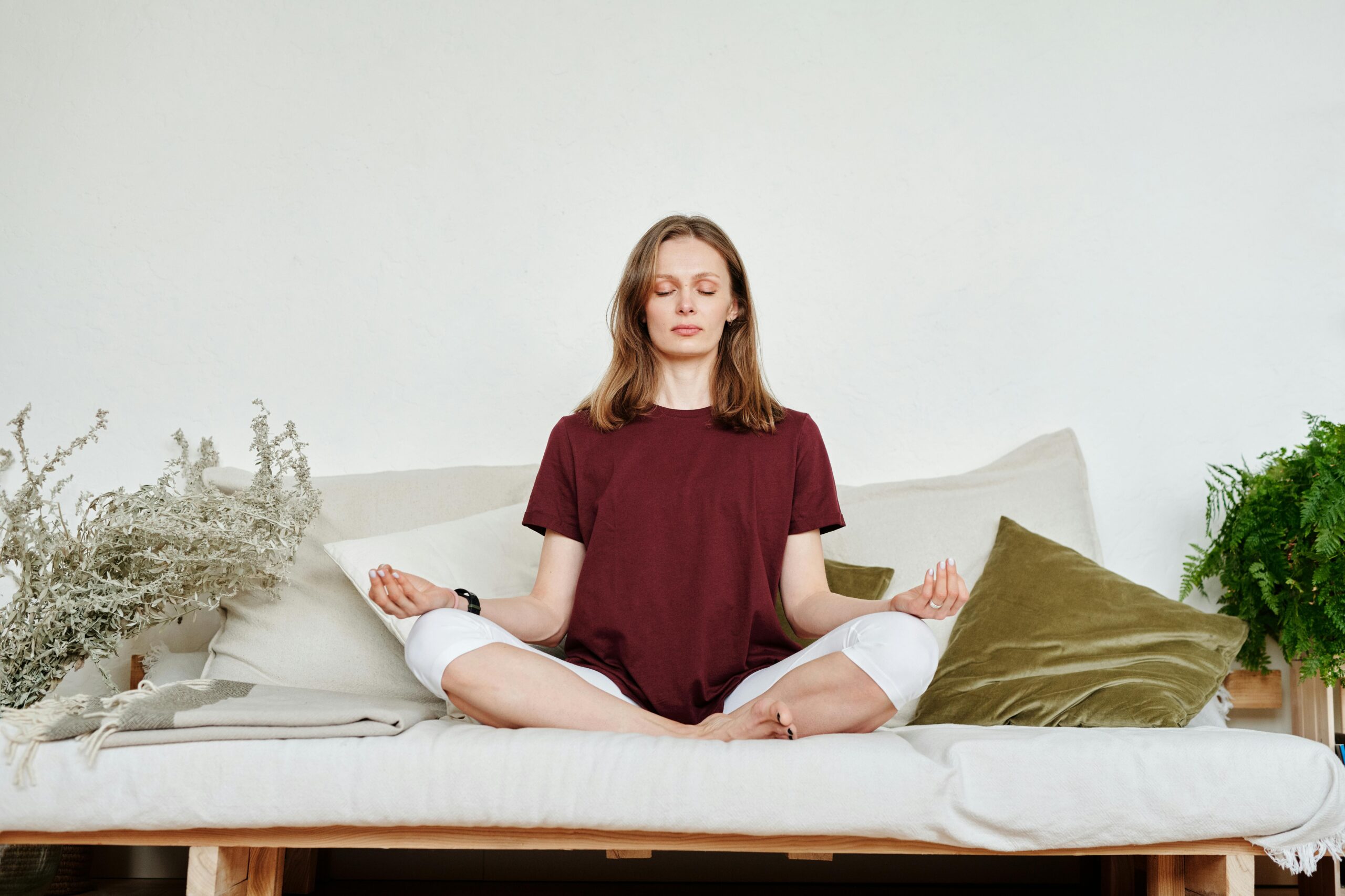 Woman with maroon short sleeve shirt sitting cross legged on a bed with white linen, a green throw pillow by her left leg with arms peacefully resting on her legs and eyes closed. There are green plants on either side of the bed.