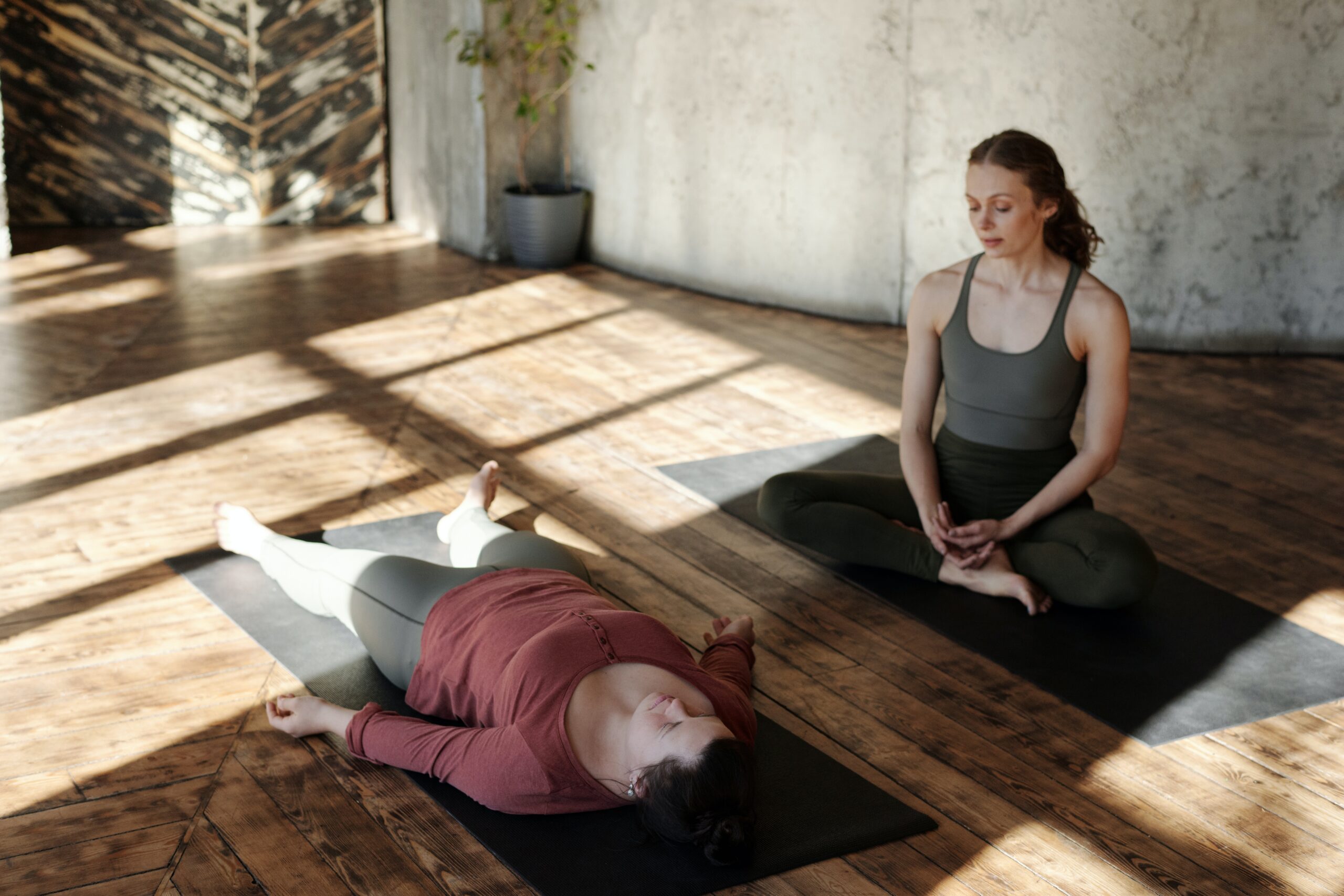 A yoga instructor and a participant on separate yoga mats with the instructor sitting cross legged and participant laying flat on her back on the mat in front of the instructor. They are in an open room with wooden floor and the sun casting a shadow over then through the framed window.