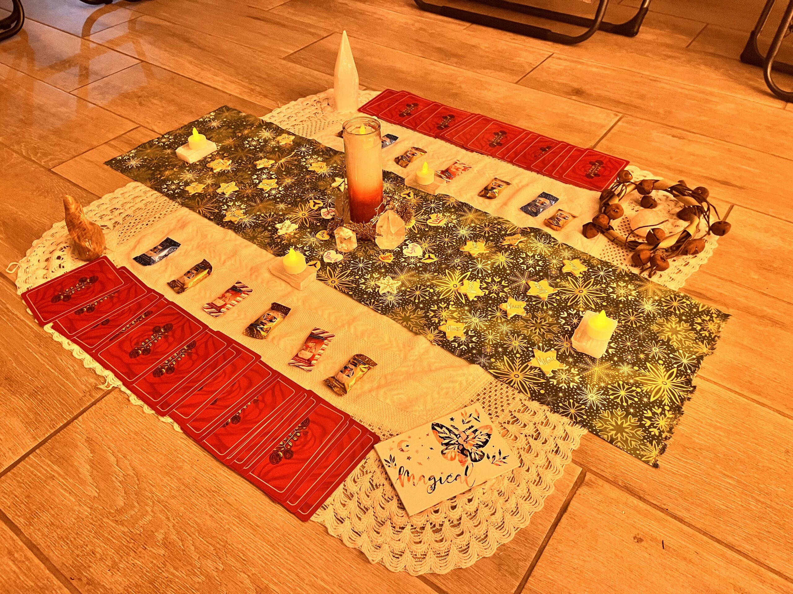 Altar dressed with a large white blanket, yuletide cloth, candles, various types of candies, crystals, oracle cards, and wooden stars with the word magic on them. This altar is set in the middle of a wooden floor.