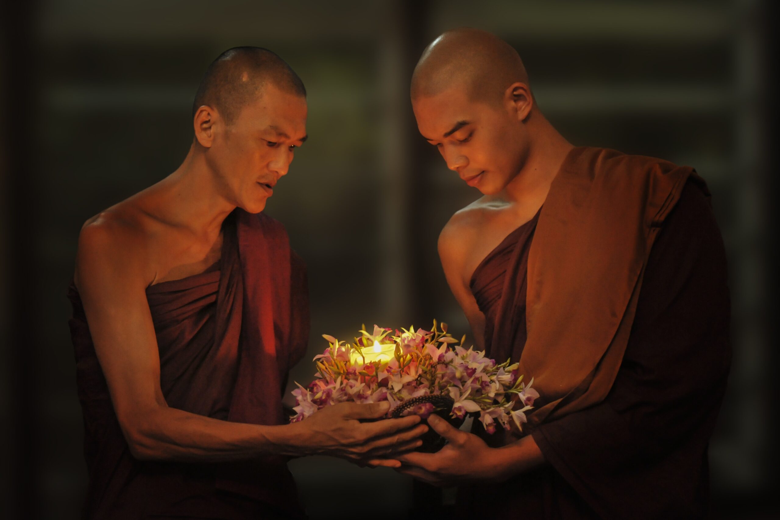 Two monks holding and looking down at a short, lit candle wrapped in brown forage in a poorly lit area, wearing a two-toned brown yoga.