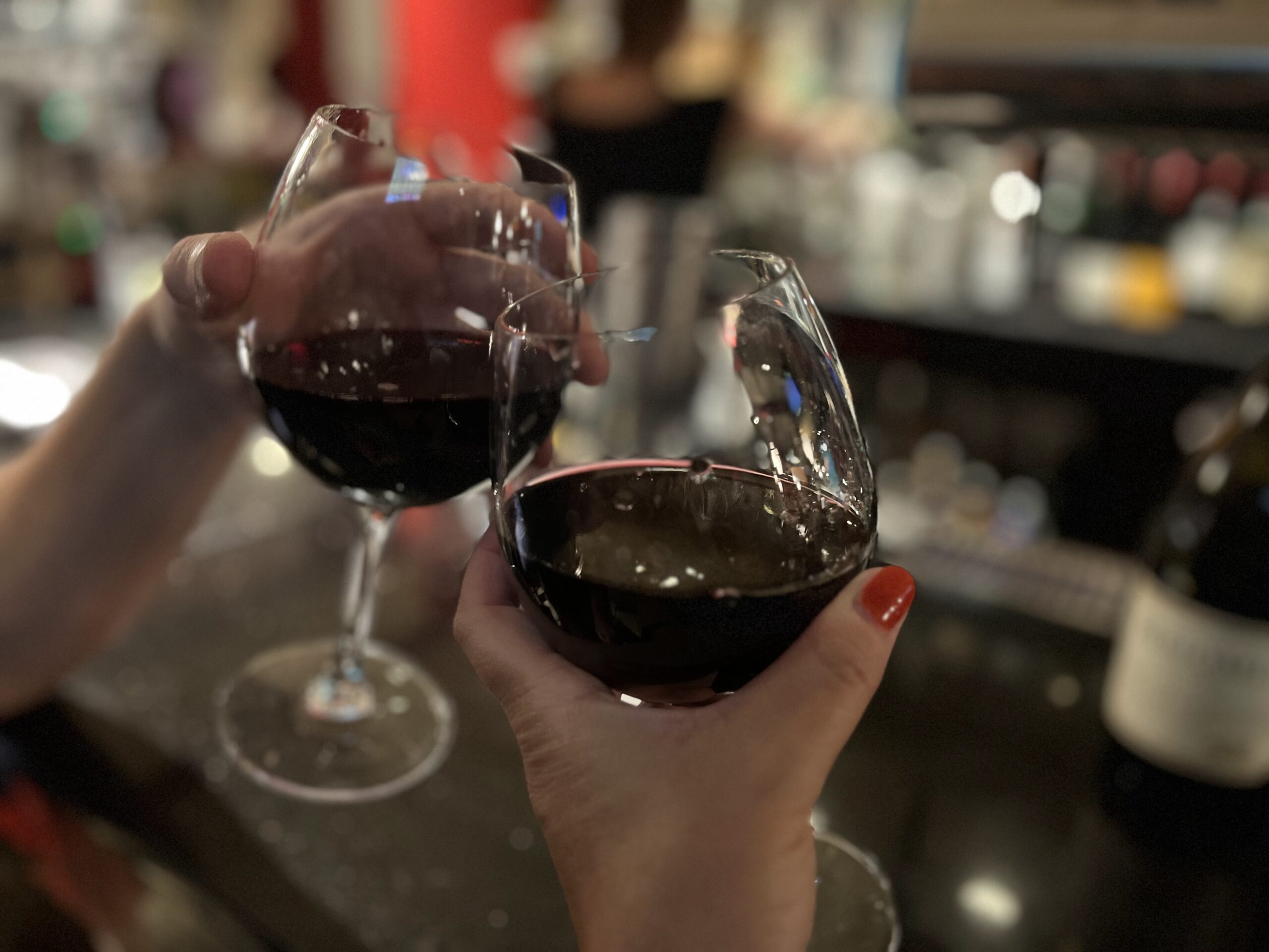 Two hands holding up two wine glasses next to each other at a bar.