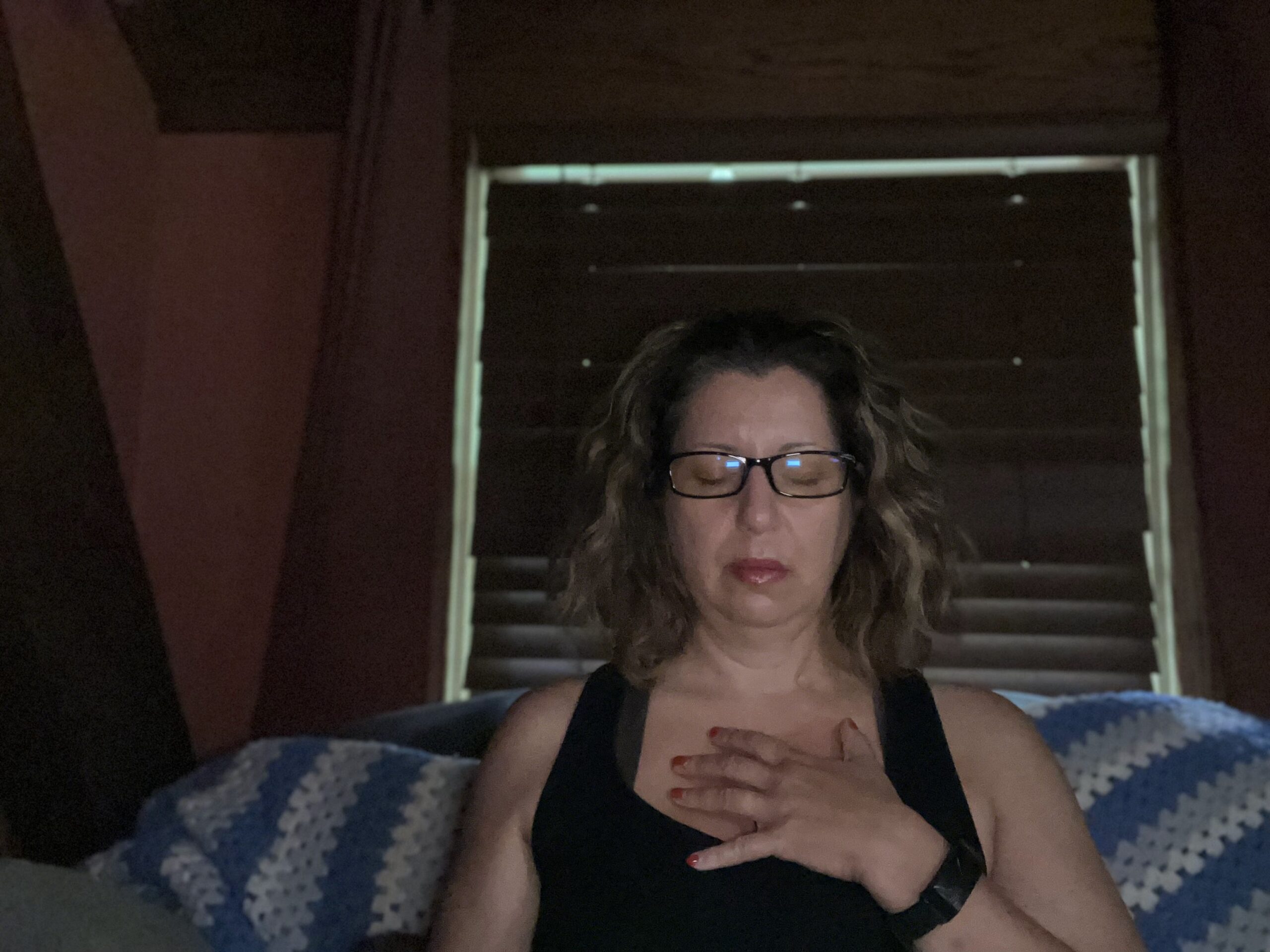 A woman sits in a lowly lit room with eyes closed and hand over her chest as she takes a moment to meditate.