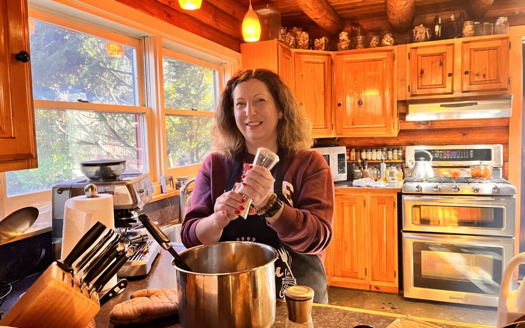 Woman standing in her kitchen holding a pepper grinder with both hands over a large pot with ladle in it. On the countertop besides her is a knife set, paper towel roll, and an espresso machine. In the background is a stove with double ovens and a countertop with racks of spices and a microwave.