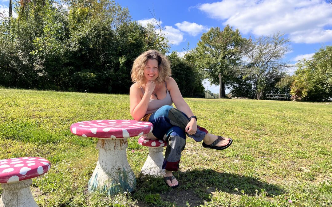 A woman smiles while sitting in an open field, on a red polka-dotted topped mushroom shaped stool, next to a table with similar colors and shape.