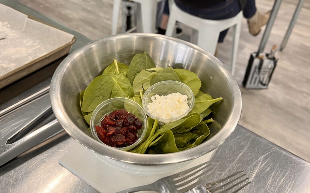 Metal bowl halfway filled with green baby spinach and two clear glass spice bowls with dried cranberries in one and cheese in the other. The bowl sits on a paper towel next to two forks which sits on metal prep table.