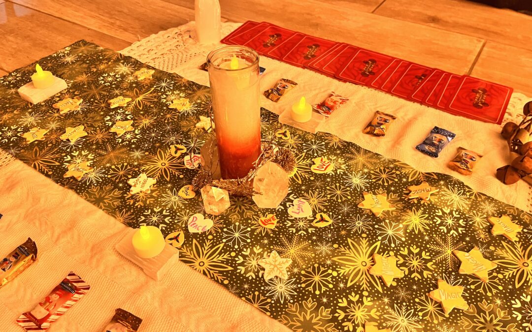 Altar dressed with a large white blanket, yuletide cloth, candles, various types of candies, crystals, oracle cards, and wooden stars with the word magic on them.